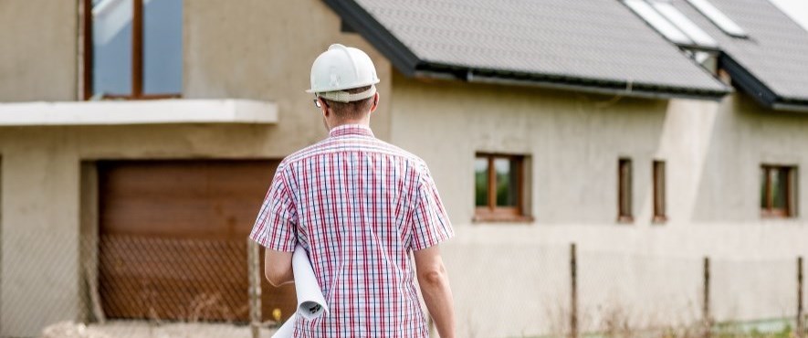 man walking towards unfinished house with plans under arm and hard hat on